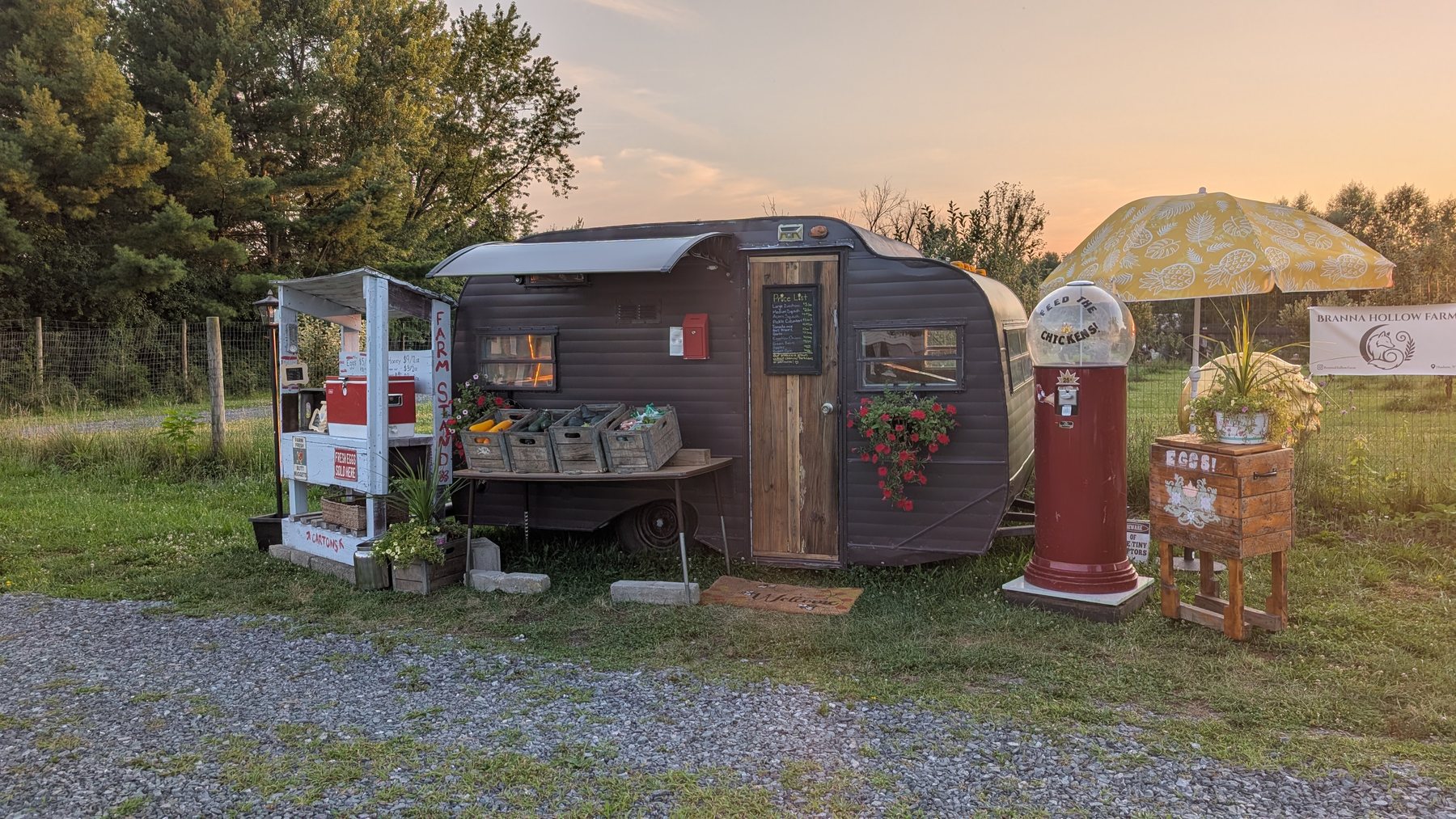 The Branna Hollow farm stand at sunset — a vintage camper with crates of produce on the table, a red gumball machine, an egg box and a yellow umbrella.